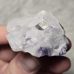 May include: A close-up of a raw fluorite crystal cluster. The crystal features a translucent, icy white exterior with a small, geometric, light-yellow and purple crystal formation. The base of the crystal has a deep purple hue.