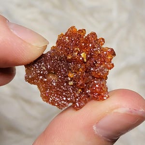 Puede incluir: Primer plano de una mano sosteniendo un grupo de pequeños cristales de color rojo anaranjado brillante. Los cristales tienen una superficie rugosa y texturizada y están agrupados. El fondo es blanco suave y borroso.