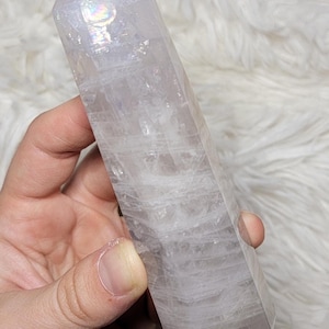 May include: A clear quartz crystal point with a polished, faceted shape. The crystal is held in a hand and has internal striations and a subtle iridescent sheen. The background is a soft, white, textured surface.