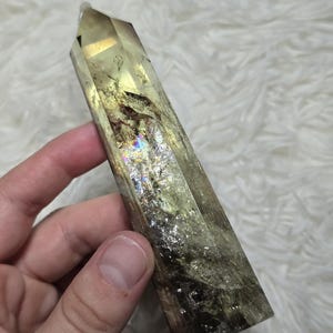 Può includere: Una torre di cristallo trasparente e sfaccettata con una parte superiore a punta. Il cristallo ha una tonalità giallo-verde con inclusioni interne e lampi iridescenti. Il cristallo è tenuto su uno sfondo bianco soffice.
