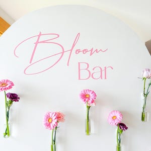 May include: A white round sign with the text "Bloom Bar" in pink lettering.  Pink flowers are arranged in clear glass tubes attached to the sign.