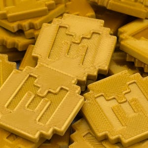May include: A pile of gold-colored, pixelated coins. Each coin has a square shape with a stylized letter "M" in the center. The coins have a textured surface and a slightly rounded edge, giving them a three-dimensional appearance.