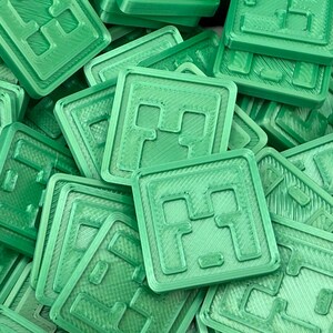 Puede incluir: Un mont&oacute;n de objetos cuadrados de color verde claro con un dise&ntilde;o en relieve. Cada cuadrado tiene un borde texturizado y un patr&oacute;n central de una cara pixelada. Los objetos parecen estar impresos en 3D y est&aacute;n dispuestos al azar.