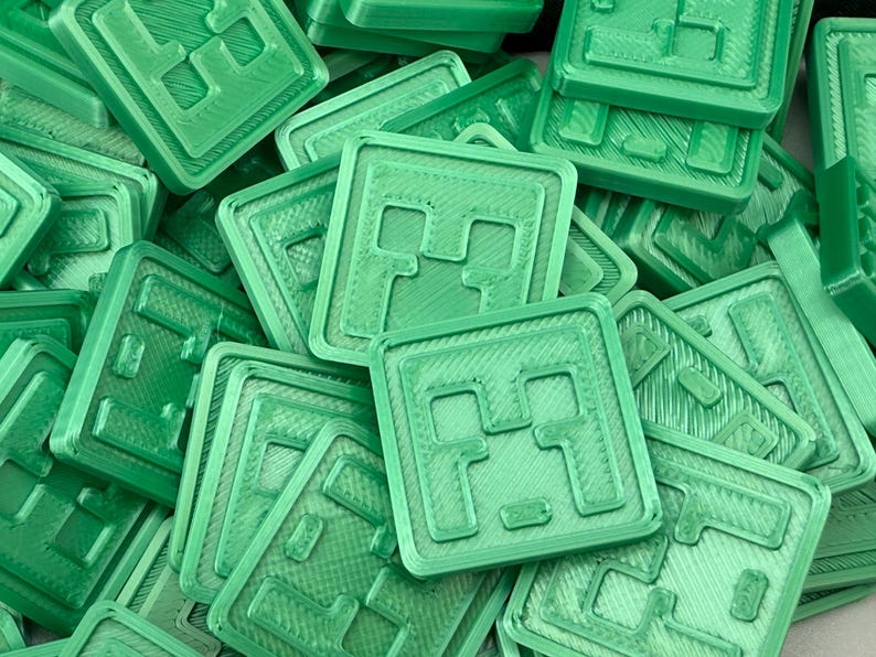 Puede incluir: Un mont&oacute;n de objetos cuadrados de color verde claro con un dise&ntilde;o en relieve. Cada cuadrado tiene un borde texturizado y un patr&oacute;n central de una cara pixelada. Los objetos parecen estar impresos en 3D y est&aacute;n dispuestos al azar.