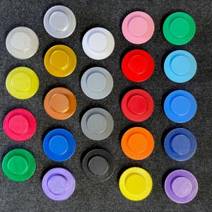 May include: A collection of twenty-four colorful, circular objects with a raised center, arranged on a dark gray surface. The objects are in various colors, including silver, gold, red, blue, and green. The objects are approximately 1.5 inches in diameter.