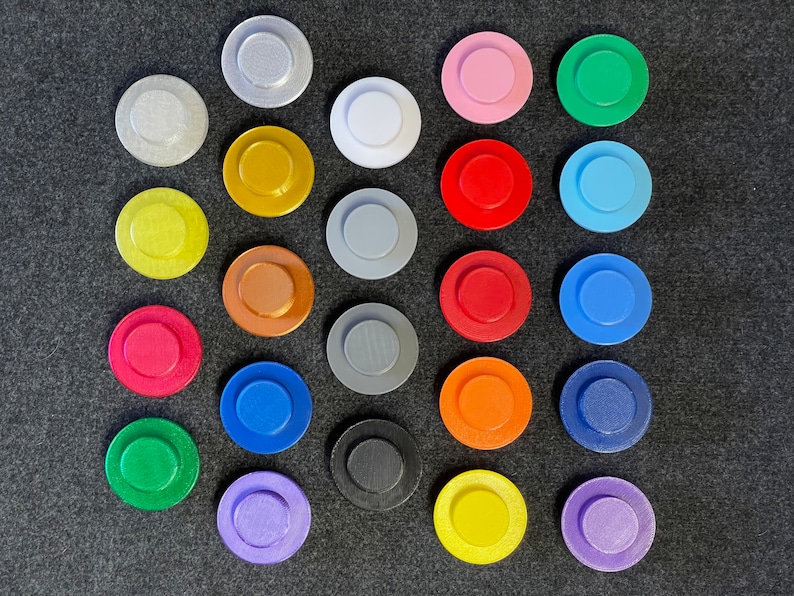 May include: A collection of twenty-four colorful, circular objects with a raised center, arranged on a dark gray surface. The objects are in various colors, including silver, gold, red, blue, and green. The objects are approximately 1.5 inches in diameter.