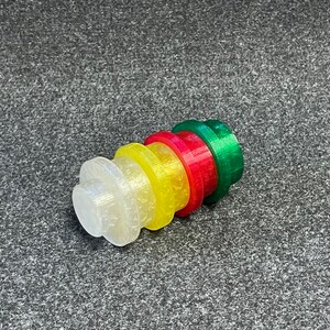 May include: A stack of colorful, 3D-printed bottle stoppers. The stoppers are in a gradient of colors: clear, yellow, red, and green. Each stopper has a circular top and a cylindrical base. The stoppers are made of plastic.