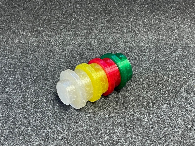 May include: A stack of colorful, 3D-printed bottle stoppers. The stoppers are in a gradient of colors: clear, yellow, red, and green. Each stopper has a circular top and a cylindrical base. The stoppers are made of plastic.