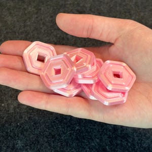 May include: A handful of pink, hexagonal tokens with a square cut-out in the center. The tokens are held in an open hand against a dark gray background. The tokens appear to be 3D printed.