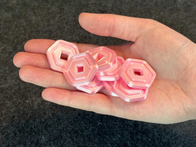 May include: A handful of pink, hexagonal tokens with a square cut-out in the center. The tokens are held in an open hand against a dark gray background. The tokens appear to be 3D printed.
