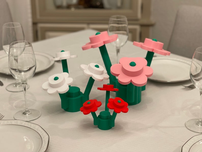 Puede incluir: Un conjunto de flores de madera estilizadas en rosa, blanco y rojo, con tallos y bases verdes. Las flores est&aacute;n dispuestas sobre un mantel blanco, con copas de vino y platos en el fondo. Un art&iacute;culo decorativo.