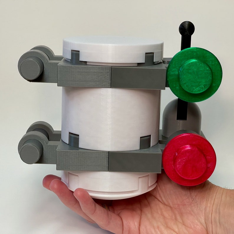 May include: A 3D-printed, cylindrical object with a white body, gray supports, and green and red circular accents. The object has a mechanical appearance, with a handle and circular buttons. The design is modern and geometric.