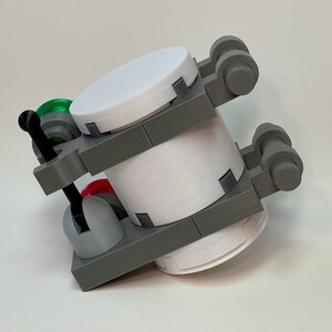 May include: A 3D-printed model of a cylindrical object with a white body and gray structural supports. It features a white lid, red and green accents, and small wheels. The model is designed with a mechanical aesthetic.