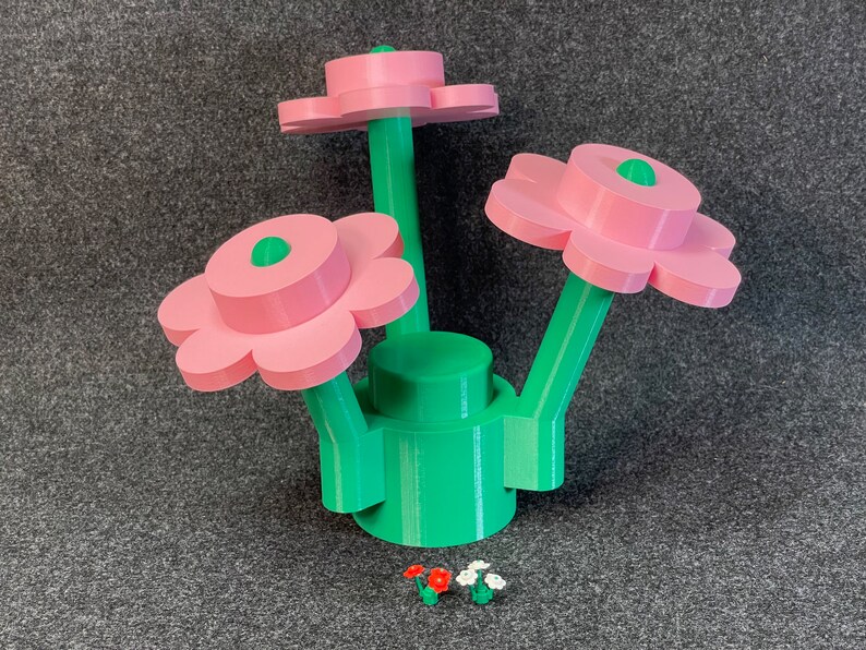 Puede incluir: Una escultura floral impresa en 3D con tres flores rosas, tallos verdes y una base verde. Las flores tienen un centro verde. Dos arreglos florales en miniatura est&aacute;n en la base de la escultura.