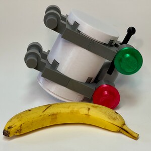 May include: A white and gray 3D-printed object with a green and red button, and a black handle. A yellow banana is in front of the object. The object has a cylindrical shape with gray brackets.