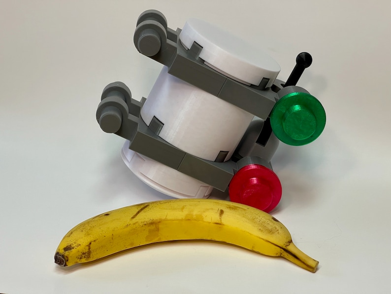 May include: A white and gray 3D-printed object with a green and red button, and a black handle. A yellow banana is in front of the object. The object has a cylindrical shape with gray brackets.