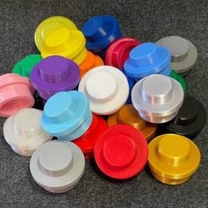 May include: A collection of colorful, 3D-printed toy building block toppers in various colors, including pink, blue, yellow, and purple. The toppers are round with a raised center, resembling oversized Lego bricks. They are arranged on a dark gray surface.