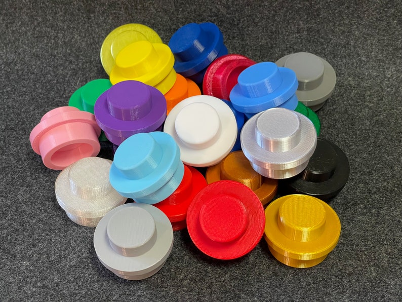 May include: A collection of colorful, 3D-printed toy building block toppers in various colors, including pink, blue, yellow, and purple. The toppers are round with a raised center, resembling oversized Lego bricks. They are arranged on a dark gray surface.