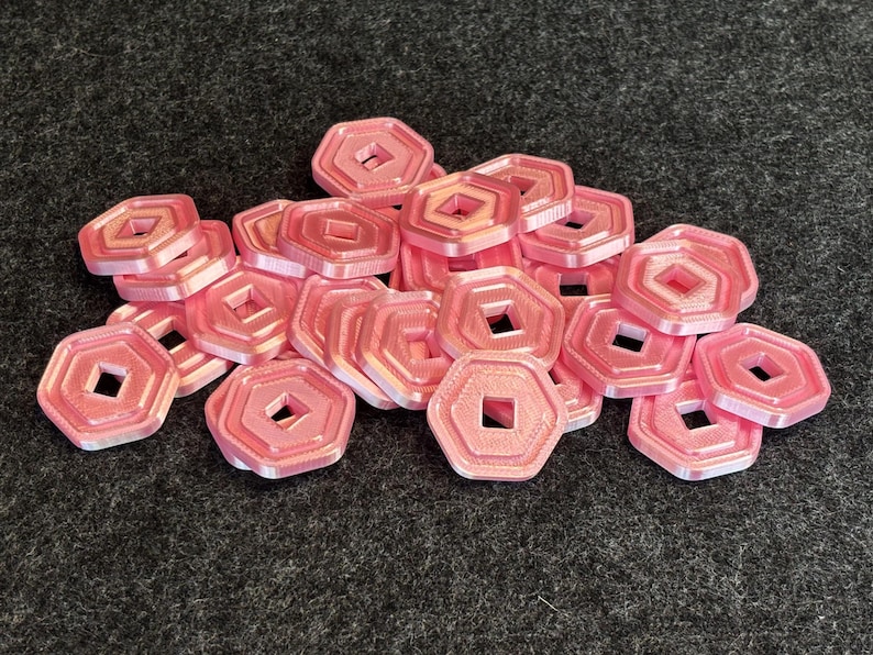 May include: A pile of pink, octagonal game tokens with a square hole in the center. The tokens are stacked on a dark gray, textured surface. The tokens have a layered design.