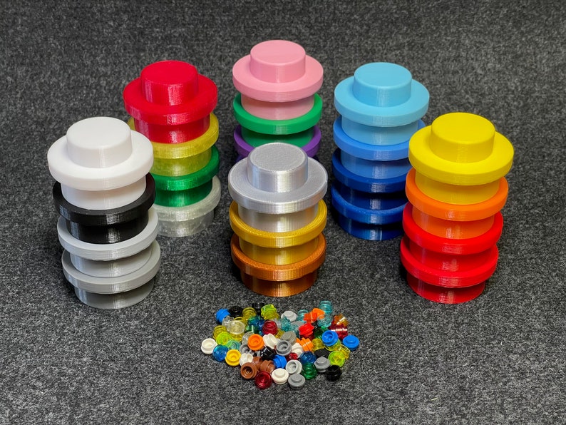 May include: A collection of colorful, 3D-printed building block accessories. Stacks of various colors, including red, pink, blue, and yellow, are arranged around a pile of small, translucent pieces. The pieces are designed to fit together.