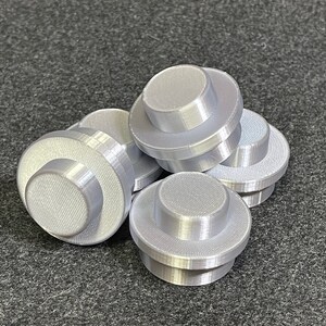 May include: Several light grey, 3D-printed, cylindrical objects with a stepped design. Each object has a circular base and a smaller, raised circular top. The objects are scattered on a dark grey surface.