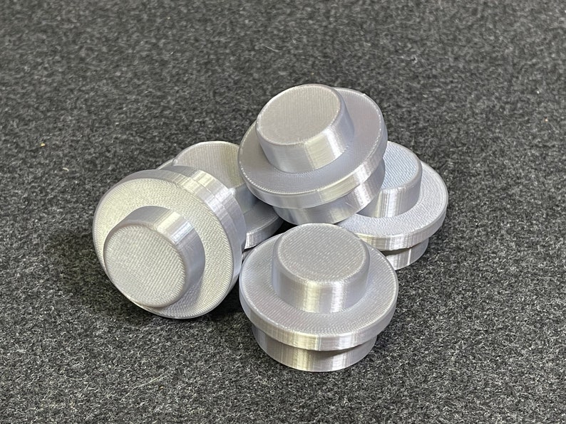 May include: Several light grey, 3D-printed, cylindrical objects with a stepped design. Each object has a circular base and a smaller, raised circular top. The objects are scattered on a dark grey surface.