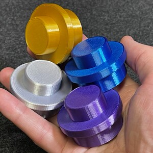 May include: Four colorful, 3D-printed objects in gold, silver, blue, and purple. Each object has a circular base and a smaller, raised circular top. The objects are held in a hand, showcasing their varied colors and shapes.