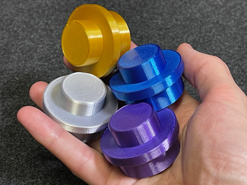 May include: Four colorful, 3D-printed objects in gold, silver, blue, and purple. Each object has a circular base and a smaller, raised circular top. The objects are held in a hand, showcasing their varied colors and shapes.