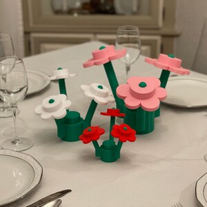 May include: A colorful floral centerpiece with green stems and bases, and flower heads in white, pink, and red. The flowers are arranged on a white tablecloth, with place settings and glassware visible in the background.