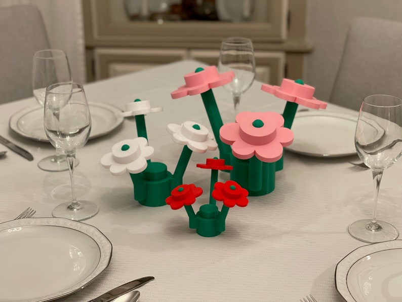 May include: A colorful floral centerpiece with green stems and bases, and flower heads in white, pink, and red. The flowers are arranged on a white tablecloth, with place settings and glassware visible in the background.