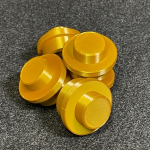 May include: A collection of six gold-coloured, circular objects with a stepped design. Each object has a flat top and a wider base, resembling a button or a small, stylised hat. The objects are arranged on a dark grey surface.