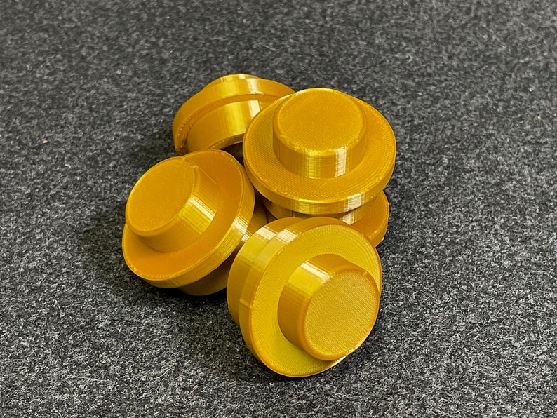 May include: A collection of six gold-coloured, circular objects with a stepped design. Each object has a flat top and a wider base, resembling a button or a small, stylised hat. The objects are arranged on a dark grey surface.