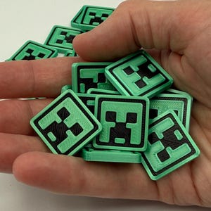 May include: A collection of square, green and black game pieces, each featuring a pixelated face design. The pieces are held in a hand, with several more scattered in the background. The design is reminiscent of a popular video game.