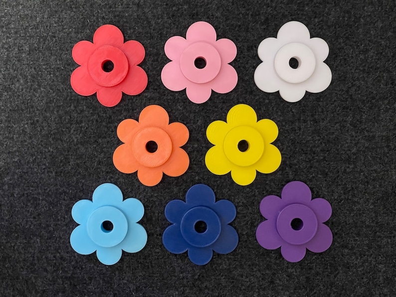 May include: A collection of seven colorful flower-shaped objects in red, pink, white, orange, yellow, blue, and purple. Each flower has a circular center and a layered design. The objects are arranged on a dark gray surface.