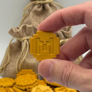 May include: A gold-colored, pixelated coin with a stylized "M" design is held between fingers. A burlap sack is in the background, with more coins scattered below. The coins appear to be game tokens or decorative items.