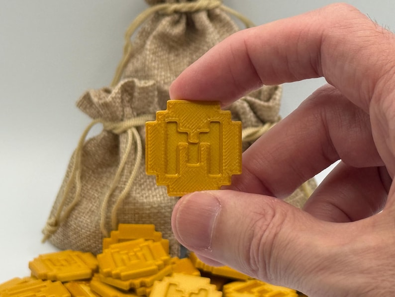 May include: A gold-colored, pixelated coin with a stylized "M" design is held between fingers. A burlap sack is in the background, with more coins scattered below. The coins appear to be game tokens or decorative items.