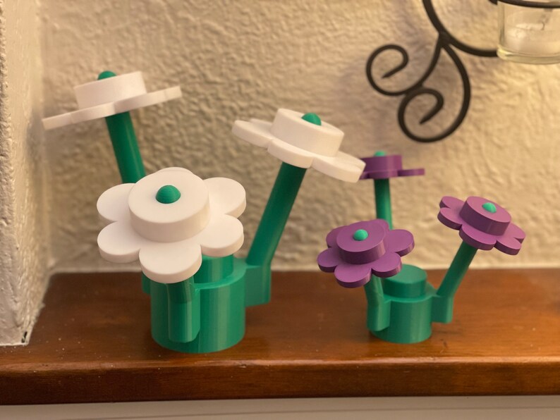 Puede incluir: Una colecci&oacute;n de esculturas de flores impresas en 3D en blanco y morado, con tallos y bases verdes. Los p&eacute;talos de las flores est&aacute;n en capas, y cada flor tiene un peque&ntilde;o centro verde y redondo. Las esculturas se exhiben sobre una superficie de madera.