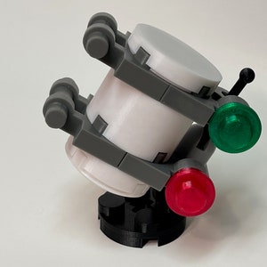 May include: A 3D-printed model of a futuristic device. The device features a white cylindrical body with gray support structures and two circular end caps. It has a black base and is adorned with a green and a red light.