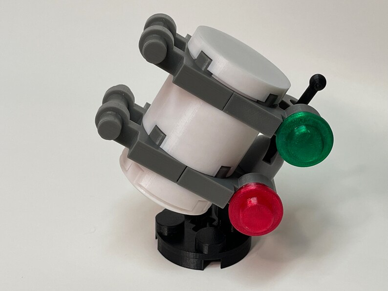 May include: A 3D-printed model of a futuristic device. The device features a white cylindrical body with gray support structures and two circular end caps. It has a black base and is adorned with a green and a red light.