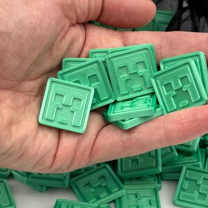 Puede incluir: Un conjunto de fichas cuadradas de color verde claro con un dise&ntilde;o de cara pixelada, sostenidas en una mano. Las fichas est&aacute;n apiladas y dispersas, lo que sugiere un juego o un objeto de colecci&oacute;n. El dise&ntilde;o recuerda a un videojuego popular.