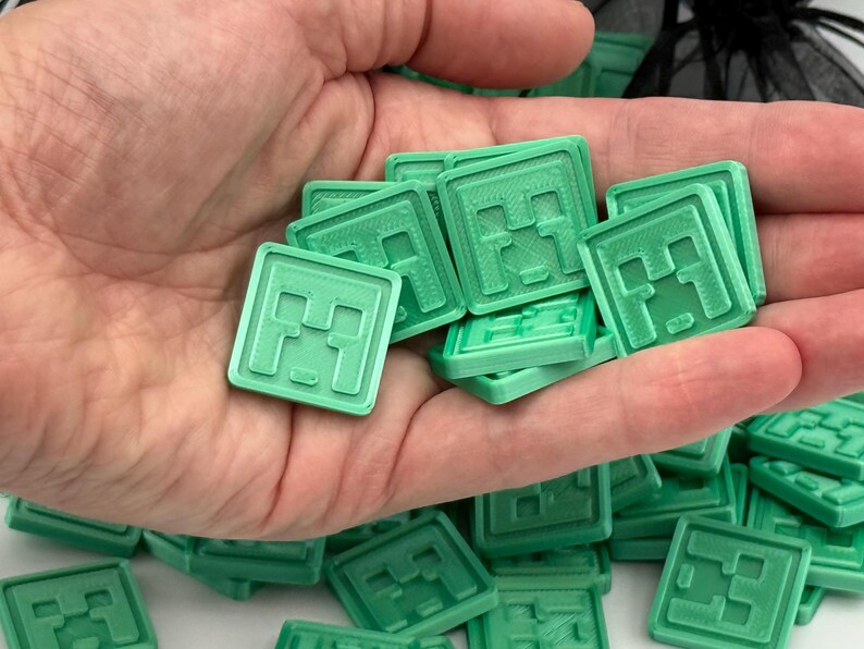 Puede incluir: Un conjunto de fichas cuadradas de color verde claro con un dise&ntilde;o de cara pixelada, sostenidas en una mano. Las fichas est&aacute;n apiladas y dispersas, lo que sugiere un juego o un objeto de colecci&oacute;n. El dise&ntilde;o recuerda a un videojuego popular.