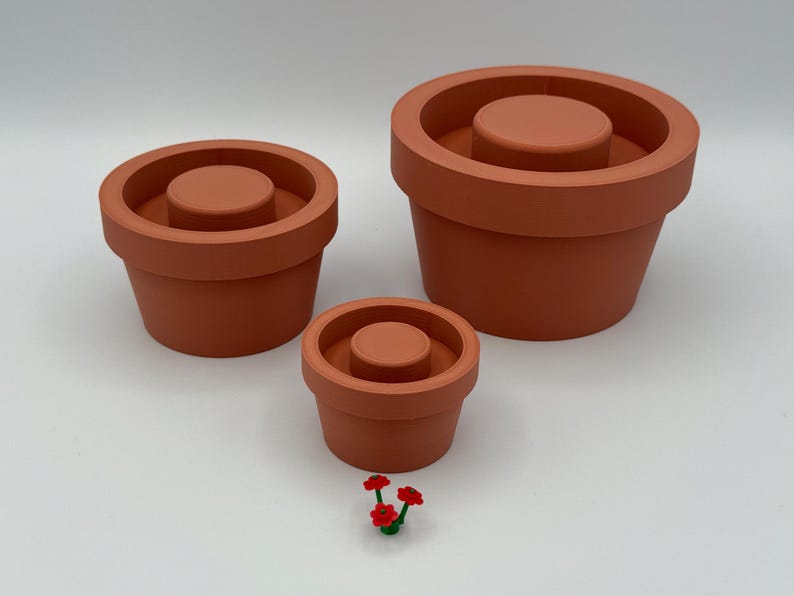 Puede incluir: Tres macetas de color terracota de diferentes tama&ntilde;os, cada una con un disco central elevado. Un peque&ntilde;o grupo de flores artificiales rojas y verdes se encuentra frente a las macetas. Las macetas est&aacute;n vac&iacute;as.