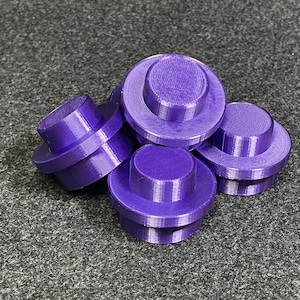 May include: A group of six purple, 3D-printed, cylindrical objects with a stepped design. Each object has a wide base and a smaller, raised top. The objects are scattered on a grey surface.