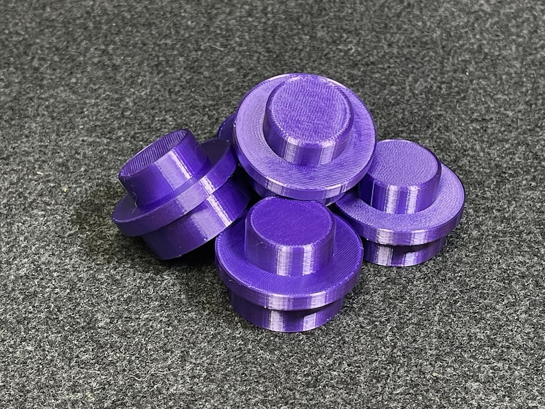 May include: A group of six purple, 3D-printed, cylindrical objects with a stepped design. Each object has a wide base and a smaller, raised top. The objects are scattered on a grey surface.