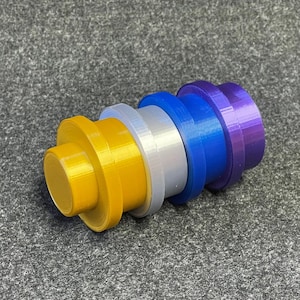 May include: A multi-coloured, cylindrical object with stacked, circular components. The colours include gold, silver, blue, and purple. The object appears to be made of plastic and is resting on a grey surface.