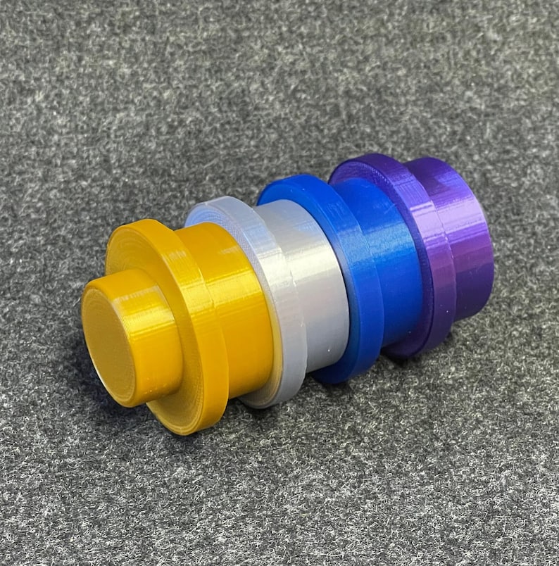 May include: A multi-coloured, cylindrical object with stacked, circular components. The colours include gold, silver, blue, and purple. The object appears to be made of plastic and is resting on a grey surface.
