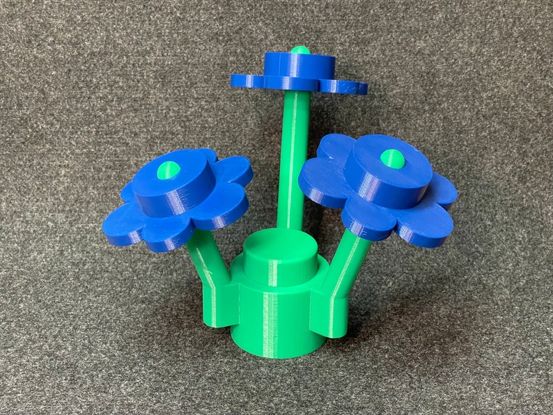 Puede incluir: Una escultura de flores impresa en 3D con tres flores azules, cada una con un centro verde. Los tallos y la base son verdes. La escultura est&aacute; sobre un fondo gris.