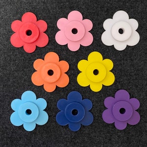 Puede incluir: Un surtido de piezas artesanales en forma de flor de colores rojo, rosa, blanco, naranja, amarillo, azul, marino y morado. Cada flor tiene un centro circular con un agujero, sobre un fondo gris oscuro.