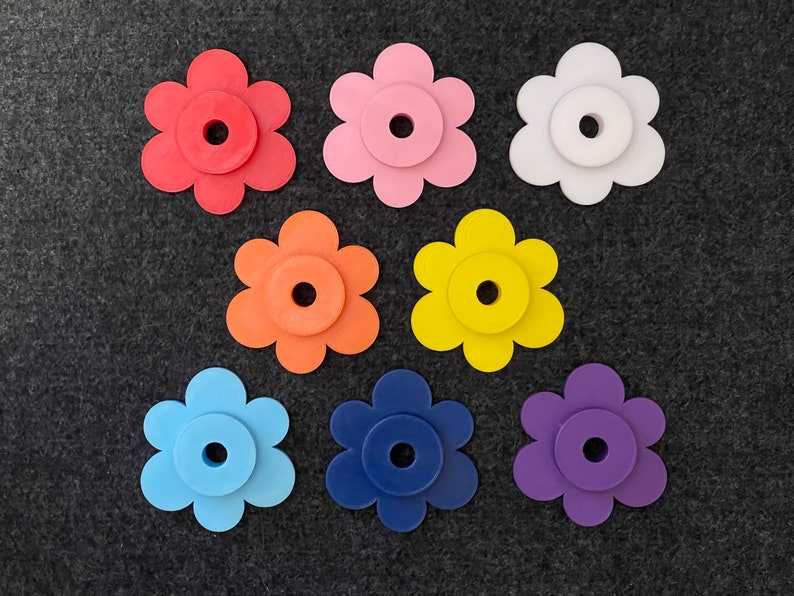 Puede incluir: Un surtido de piezas artesanales en forma de flor de colores rojo, rosa, blanco, naranja, amarillo, azul, marino y morado. Cada flor tiene un centro circular con un agujero, sobre un fondo gris oscuro.