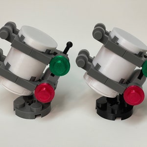 May include: Two miniature, futuristic-looking models. Each has a white cylindrical body, gray supports, and a base. One has a red and green light, the other has a red and green light. The bases are gray and black.